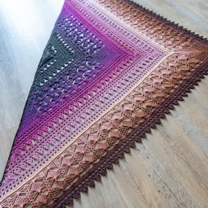 Può includere: Uno scialle all'uncinetto in una sfumatura di colori, che passa dal viola scuro al marrone chiaro. Lo scialle presenta intricati motivi traforati e un bordo smerlato. L'accessorio fatto a mano è esposto su una superficie di legno.