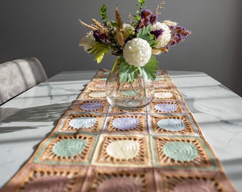 May include: A colorful, crocheted table runner with a repeating square pattern in shades of brown, purple, green, and cream. A clear glass vase holds a bouquet of white and purple flowers, adding a touch of elegance to the table setting.