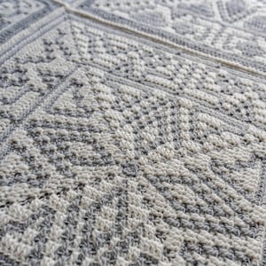 May include: Close-up of a crocheted blanket with a geometric pattern. The blanket features a combination of white and grey yarn, creating a textured surface. The design includes various shapes and patterns, showcasing the craftsmanship of the handmade item.