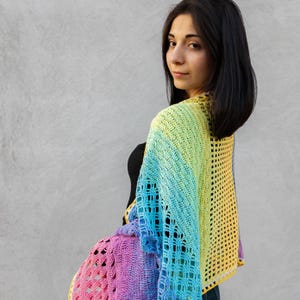 May include: A woman wearing a rainbow crochet shawl with a yellow, blue, green, and purple color gradient. The shawl is crocheted in a delicate openwork pattern.