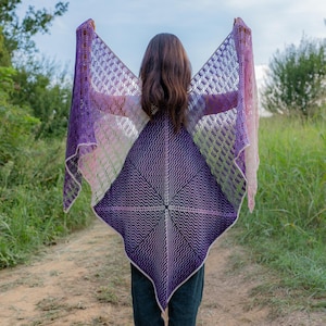 Puede incluir: Un chal de ganchillo morado y blanco con un patrón geométrico. El chal está siendo sostenido por una persona, mostrando la parte posterior del chal.