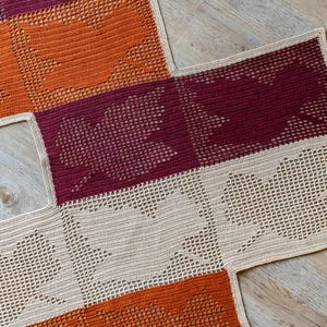 May include: A crocheted blanket with squares in orange, burgundy, and beige. The blanket is made with a lacy stitch pattern.