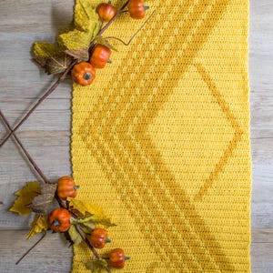 May include: A yellow crocheted table runner with a diamond pattern. The runner is decorated with a branch of fall leaves and small orange pumpkins.