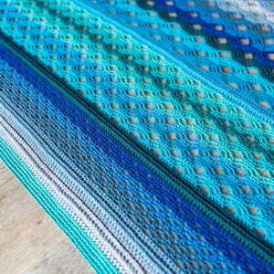 Puede incluir: Una manta de ganchillo con un patrón degradado azul, verde y gris. La manta está hecha con un punto delicado y abierto y tiene un tacto suave y texturizado.