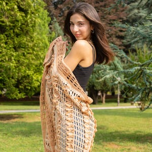 May include: A beige, open-weave crochet cardigan with a ruffled trim. The cardigan is draped over the shoulders. The background features green trees and grass.