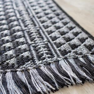 May include: Close-up of a hand-crocheted rug with a geometric pattern in shades of gray and black. The rug features a textured design with a fringe along the edge. The rug is made of yarn.