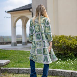 May include: A long, open-front crochet cardigan in a patchwork design. The cardigan features squares of light green, sage green, and gray yarn. The sleeves are three-quarter length. The person is wearing blue jeans.