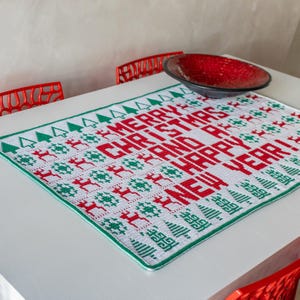 May include: White crochet table runner with a green and red Christmas design. The design features the words "Merry Christmas and Happy New Year" with small trees and reindeer.