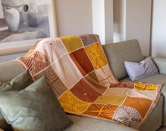Puede incluir: Una manta de ganchillo multicolor con un patrón geométrico en tonos marrones, beige y amarillos. La manta está colocada sobre un sofá con almohadas.