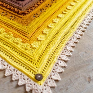 May include: A yellow crochet shawl with a white crocheted border and a brown button. The shawl is made with a gradient of yellow yarn, starting with a darker yellow at the top and fading to a lighter yellow at the bottom.