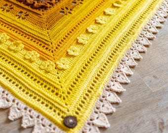 May include: A yellow crochet shawl with a white crocheted border and a brown button. The shawl is made with a gradient of yellow yarn, starting with a darker yellow at the top and fading to a lighter yellow at the bottom.