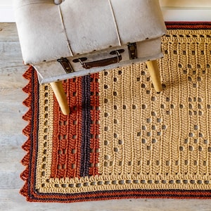 May include: A crocheted rug with a brown, orange, and beige color scheme. The rug has a geometric pattern and a fringe border. A wooden stool with a light brown fabric top sits on the rug.