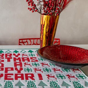 May include: A red and green Christmas table runner with the words "Merry Christmas Happy New Year!" in red. A red mosaic bowl and a vase with red berries and white flowers are on the table runner.
