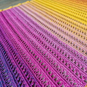 Puede incluir: Un chal de ganchillo hecho a mano en un degradado de colores, que transiciona del amarillo al rosa y al morado. El chal presenta un patrón calado detallado con rayas horizontales, creando un diseño texturizado. Los bordes están acabados con un borde decorativo.