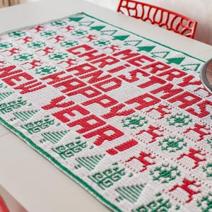 May include: A white crocheted table runner with a green and red Christmas design. The runner features the text "Merry Christmas and Happy New Year!" in red.