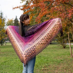 May include: A handmade, open-weave shawl in a gradient of colors, from dark purple to light peach. The shawl has a triangular shape with a decorative, scalloped edge. The background shows a park with trees and green grass.