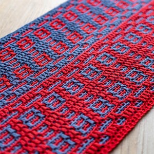 Op de afbeelding: Rood en blauw gehaakt textiel met een herhalend patroon. De stof is geweven in een strak, dicht patroon.