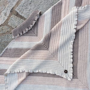 Può includere: Una coperta all'uncinetto con un motivo a righe in tonalità beige e bianco. La coperta ha un bordo a festoni e un dettaglio con un bottone.