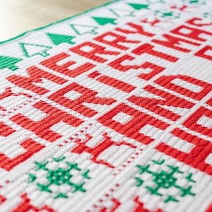 May include: White crochet blanket with red lettering that says "Merry Christmas". The blanket has a green border with small green Christmas trees and snowflakes.