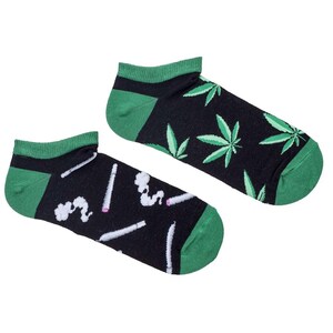 Puede incluir: Un par de calcetines negros de tobillo con ribete verde. Un calcetín tiene un patrón de hojas verdes de marihuana, mientras que el otro tiene un patrón de porros blancos de marihuana con humo.