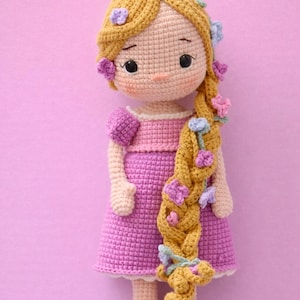 Puede incluir: Una muñeca de ganchillo hecha a mano con pelo largo y dorado adornado con flores moradas y azules. La muñeca lleva un vestido y zapatos rosas, con un corpiño rosa a juego. La cara de la muñeca tiene ojos negros, una nariz pequeña y mejillas rosadas.