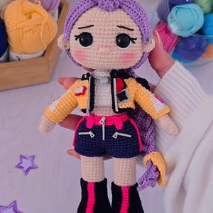 May include: Handmade crochet doll with purple hair in a bun and braid. The doll is dressed in a yellow and black jacket, navy shorts with pink trim, and black boots with pink accents. The doll is a craft item.