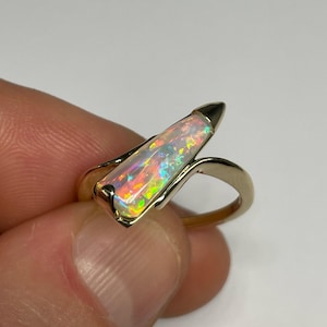 May include: A gold ring with a rectangular opal displaying iridescent colours. The opal is set in a gold band with a distinctive, curved design. The ring is held by a person's fingers.