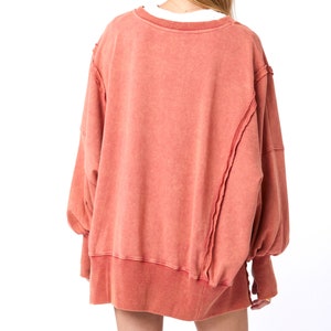 May include: A loose-fitting, long-sleeved, coral-colored sweatshirt with a distressed, unfinished hem and seams.