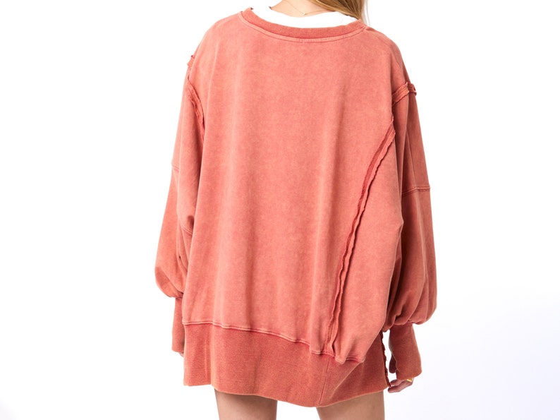 May include: A loose-fitting, long-sleeved, coral-colored sweatshirt with a distressed, unfinished hem and seams.