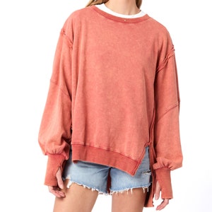 May include: A terracotta colored, oversized sweatshirt with a relaxed fit and a high-low hemline. The sweatshirt has long sleeves with thumbholes and a distressed, raw-edge detail.