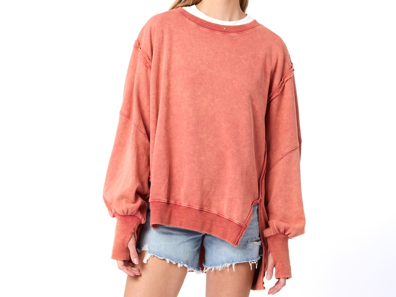 May include: A terracotta colored, oversized sweatshirt with a relaxed fit and a high-low hemline. The sweatshirt has long sleeves with thumbholes and a distressed, raw-edge detail.