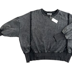 May include: A dark gray, oversized sweatshirt with a distressed look. The sweatshirt has a round neckline and long sleeves with a gathered detail at the shoulders. The sweatshirt has a raw edge detail at the hem and sides.