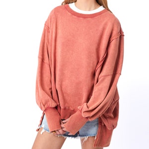 May include: A loose-fitting, oversized, salmon-colored sweatshirt with long sleeves and a crew neck. The sweatshirt has a distressed, vintage look with frayed edges and a faded appearance.