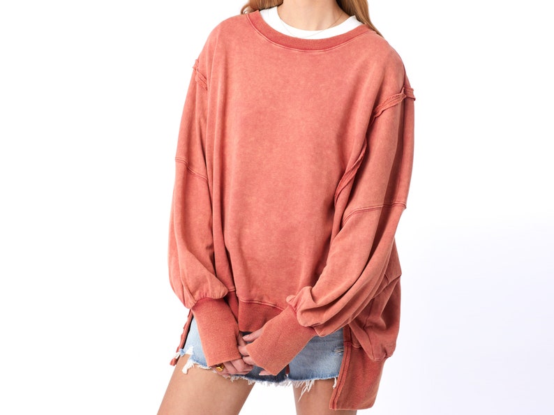 May include: A loose-fitting, oversized, salmon-colored sweatshirt with long sleeves and a crew neck. The sweatshirt has a distressed, vintage look with frayed edges and a faded appearance.