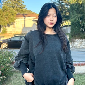 May include: A dark gray oversized sweatshirt with long sleeves and a relaxed fit. The sweatshirt has a distressed look and a slightly frayed hem.