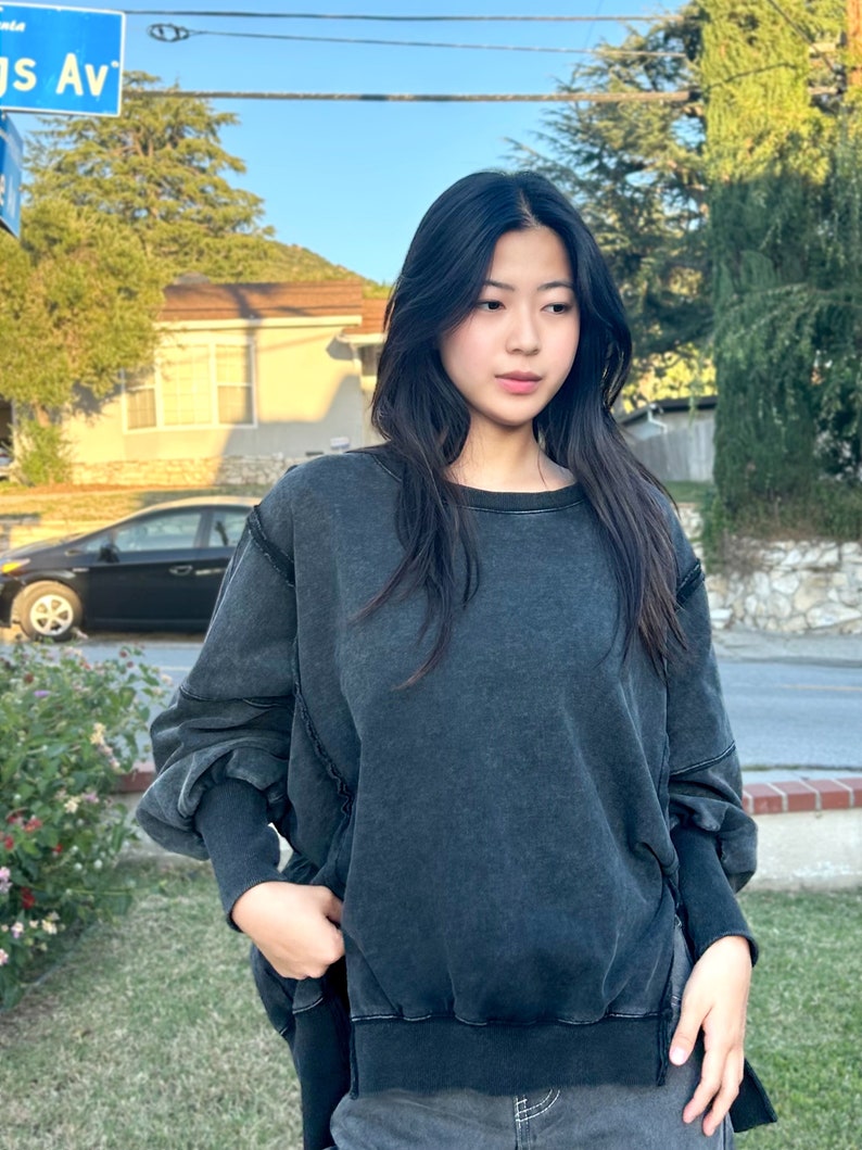 May include: A dark gray oversized sweatshirt with long sleeves and a relaxed fit. The sweatshirt has a distressed look and a slightly frayed hem.