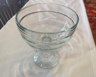 May include: A clear glass trifle bowl with a pedestal base. The bowl has a wide, open top and a decorative, ribbed design around the sides. The bowl is sitting on a white tablecloth.