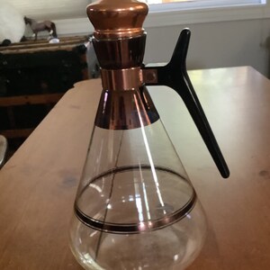 May include: A clear glass decanter with a copper top and black handle. The decanter has a gold band around the middle.