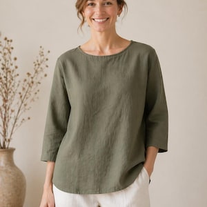 Könnte beinhalten: Ein olivgrünes Leinenoberteil mit Dreiviertelärmeln und einem U-Boot-Ausschnitt. Das Oberteil wird mit einer weißen Hose kombiniert. Die Frau lächelt. Der Hintergrund ist neutral gefärbt.