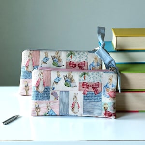 May include: Two rectangular zippered pouches with a patchwork pattern featuring rabbits, carrots, and bows in pink, blue, and white. The pouches have a light blue ribbon attached to the zipper. A pen and a stack of books are in the background.