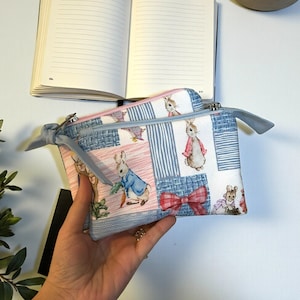 Può includere: Due piccole pochette rettangolari con cerniera con un design patchwork con illustrazioni di conigli in vari abiti. Le pochette hanno una cerniera azzurra e un nastro azzurro attaccato alla cerniera. Lo sfondo include un taccuino aperto.