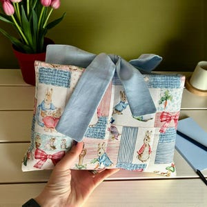 Puede incluir: Una bolsa de tela rectangular con un estampado de Peter Rabbit sobre cuadrados estilo patchwork en azul, rosa y blanco, acentuada por un gran lazo de tela azul claro liso atado en el centro. El patrón incluye conejos, zanahorias y lazos.
