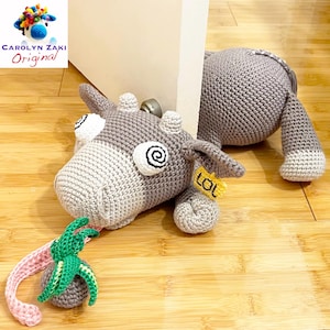 May include: A gray crocheted cow doorstop with a pink and green palm tree hanging from its mouth. The cow has a yellow tag that says "LOL".