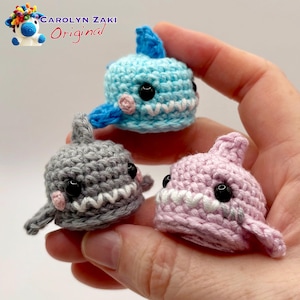 Può includere: Tre giocattoli a forma di squalo all'uncinetto fatti a mano in azzurro, grigio e rosa. Ogni squalo ha denti bianchi, occhi neri e piccoli accenti rosa. L'immagine include il testo "Carolyn Zaki Original".