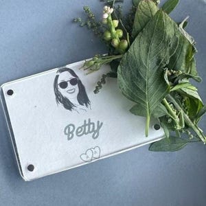 May include: A white acrylic place card with a black and white illustration of a person wearing sunglasses. The name "Betty" is written in a cursive font below the illustration. Two small hearts are printed below the name.
