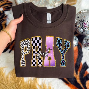 May include: A brown sweatshirt with the word "PRAY" in a multi-patterned design, including leopard print, checkered, and a purple gradient. The word "just" is written in cursive below the word "PRAY".