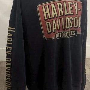 Puede incluir: Sudadera negra de Harley-Davidson con un gran logotipo rectangular en la parte delantera. El logotipo tiene un esquema de color rojo y beige y presenta el nombre Harley-Davidson y la palabra "Motorcycles". El nombre Harley-Davidson también está impreso en la manga izquierda en beige.