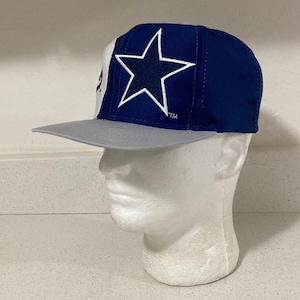 Könnte beinhalten: Eine blaue und weiße Baseballkappe mit einem großen weißen Stern auf der Vorderseite. Der Stern hat einen blauen Umriss und eine dunkelblaue Mitte. Die Kappe hat einen grauen Schirm und der Text "TM" ist auf der Vorderseite der Kappe sichtbar.