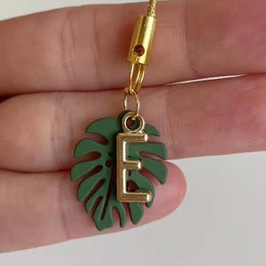 May include: A gold-colored charm with a green leaf and the letter "E". The leaf is a dark green color and has a gold-colored letter "E" attached. The charm is attached to a gold-colored cord.