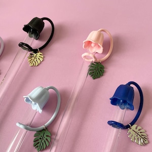 May include: A collection of colorful, decorative drink stirrers. Each stirrer features a flower-shaped top in various colors, including pink, blue, and green, with a leaf charm. The stirrers are arranged on a pink background.